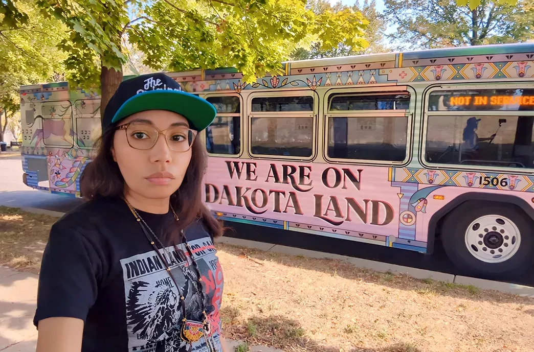 Marlena Myles in front of a bus with the words "We are on Dakota land" painted on the exterior